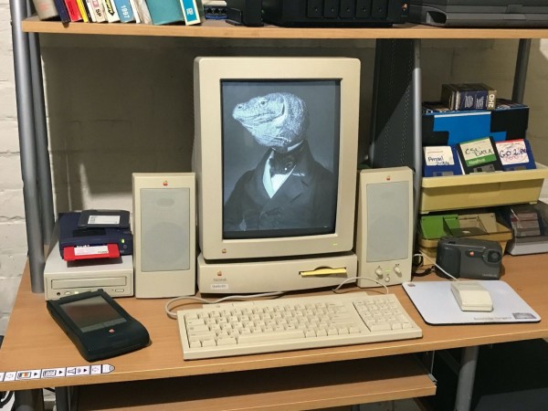 A photo of a desk with a retro mac setup on it, including a portrait monitor. On the portrait monitor is a greyscale painting in a ~17th century style of a gentlemanly monitor lizard - a monitor portrait.