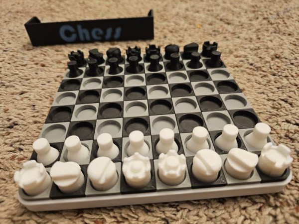 Picture of a 3d printed chessboard, with black and grey squares with little holes that the pieces fit into so they don't slide around. While and black armies are in their starting positions. In the background the strap says "Chess" in blue glow in the dark letters. 