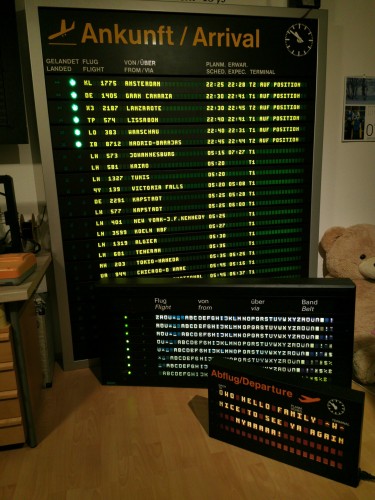Three airport information displays in varying sizes. In the back is a very tall (around two meters) board with 24 lines showing actual arrivals at my local airport. On the floor, in front of it, is a smaller board (around 60 cm tall) with 8 lines, showing the alphabet as a test text. Slightly to the right and in front of that is yet another, smaller one, about 40 cm tall, with 4 lines, showing "OWO HELLO FAMILY ^W^ NICE TO SEE YOU AGAIN NYAAAAA!" 