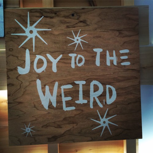 Hand-painted stars and lettering in white on plywood. The message reads “Joy to the Weird”