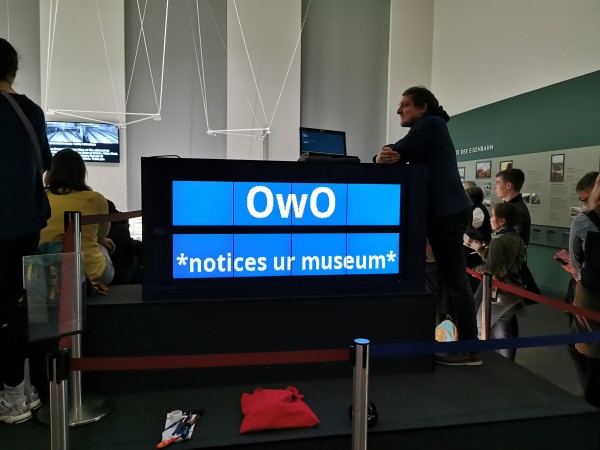 Display in the museum with people around and one person leaning on it. It reads "OwO *notices ur museum*" 
