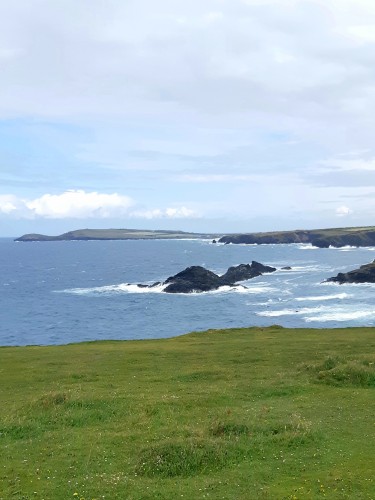 A view across the curve of a bay. Sunshine on grassy cliff tops. Rocks in the sea, with waves crashing against them. The sky blue with billowing clouds.