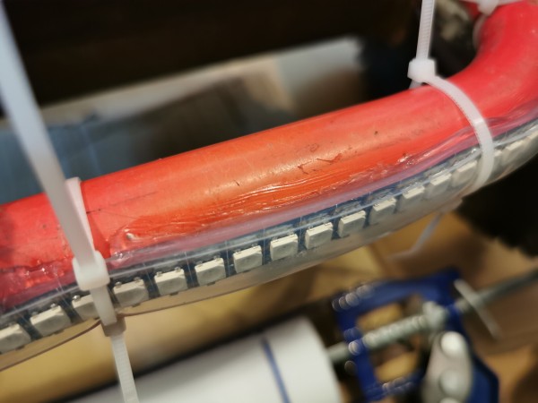 Close up of an LED strip inside a rectangular silicone tube, held with cable ties to a red part of the kart frame. Silicone is smeared between the silicone tube and the frame. 