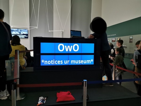 Display in the museum with people around and one person leaning on it. It reads "OwO *notices ur museum*" 