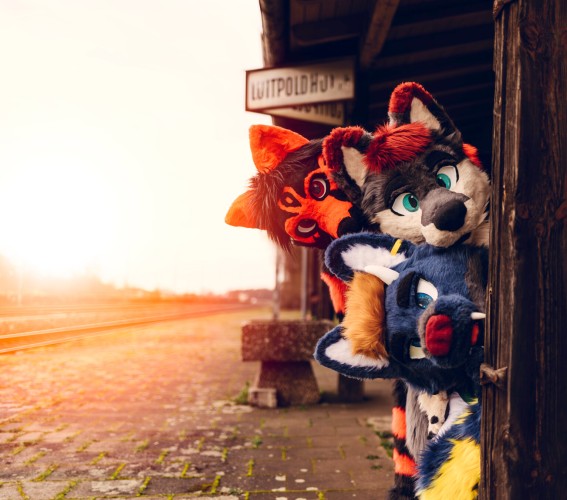 A picture of 3 Fursuiters tightly looking around a wooden support beam. The structure is standing on an abandoned railway platform, with moss growing on the rocks. In the background, typical infrastructure for a train station, including the name sign reading "Luitpoldhütte"