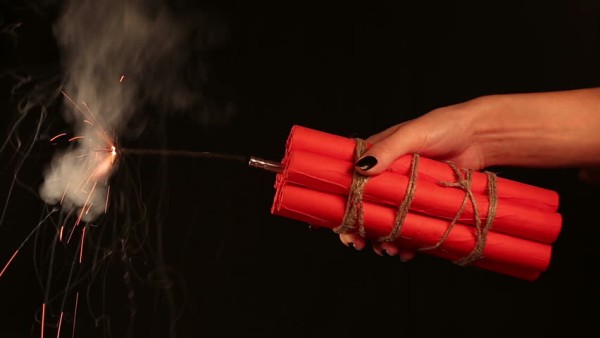 A hand holding a bundle of dynamite with the attached fuse cord burning 