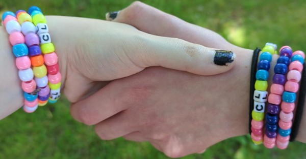 Two hands holding each other horizontally above a blurred grassy background. Both have chipped black nail polish with faint glittery accents, and each has a set of three kandi/bead bracelets on each wrist. The left hand's bracelets, moving up the wrist, have beads in the colors of the MAP flag, transage flag, and transgender flag, while the right ones have the colors of the MAP flag, bisexual flag, and transgender flag. Each MAP flag bracelet has white beads with the black letters "CL" where the MAP flag's plain white stripe would normally be. The right wrist's bracelet also has a pair of plain black bands around it above and below the bracelets.