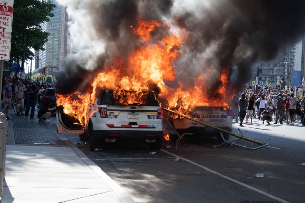 Photograph: two cop SUVs, side by side, thoroughly engulfed in orange flames which are billowing dark smoke above a large crowd of protesters beyond the cars. The day is otherwise sunny and most people in the photo appear to be facing away from the cop car bonfire