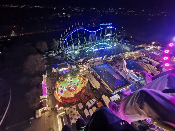 View from around 60 meters above Frankfurt at night. Part of a travelling funfair is visible, including a roller coaster lit up blue, a rotation flat ride and a large tent. On the right edge are some pink lights from the ride I'm on